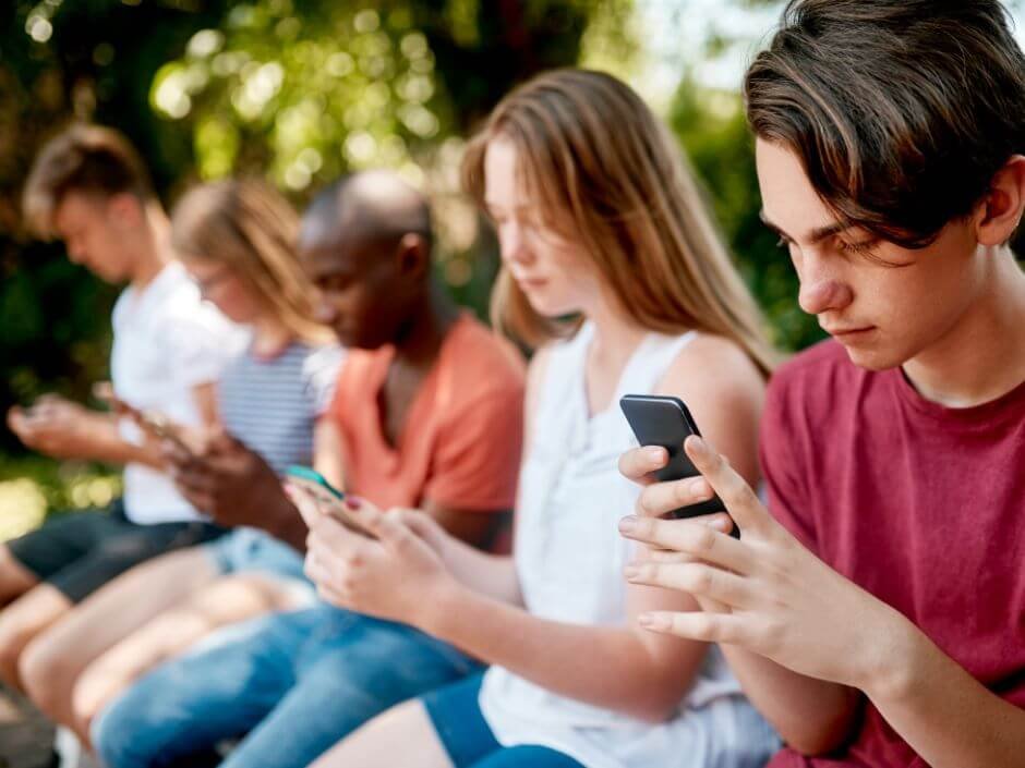 Een groep leerlingen zit op een rij buiten en kijkt op hun telefoons terwijl ze zoeken naar aanwijzingen voor de citygame City Detective.